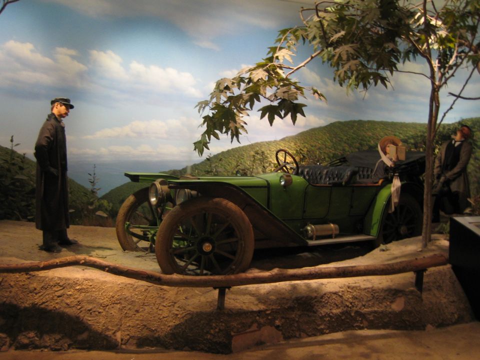 Mannequins stand near a green touring car in a museum diorama with a painted landscape background
