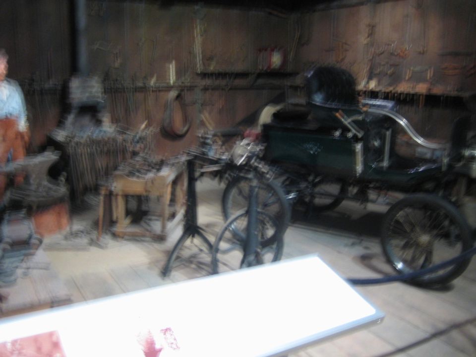 Antique green automobile with large spoked wheels and a black top, viewed from the side in a workshop setting