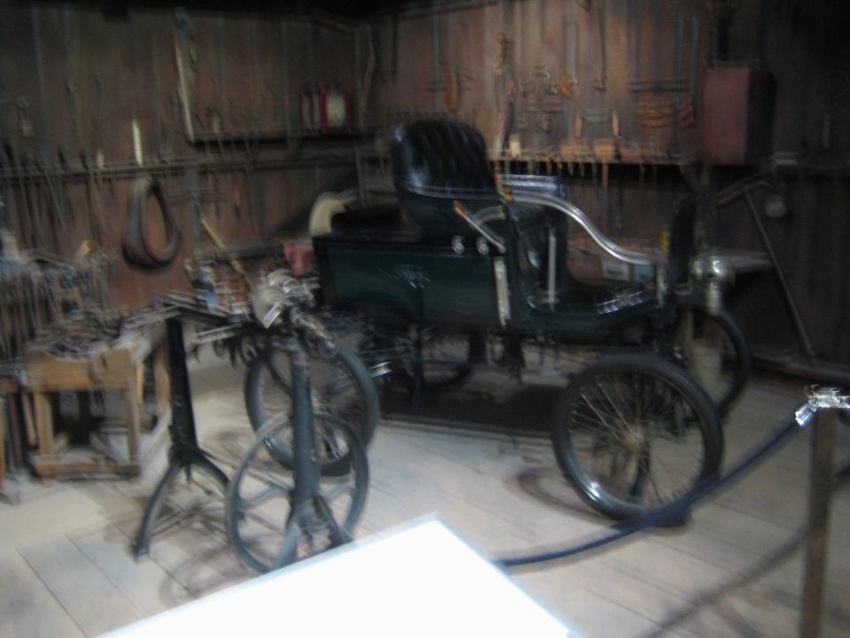 Antique horseless carriage with black leather seats and spoked wheels, parked in a workshop