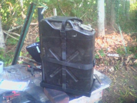 1993 Ford Bronco - A finished shop-welded steel jerry can carrier sitting on a workbench outdoors, holding a black military-style fuel can secured with black…
