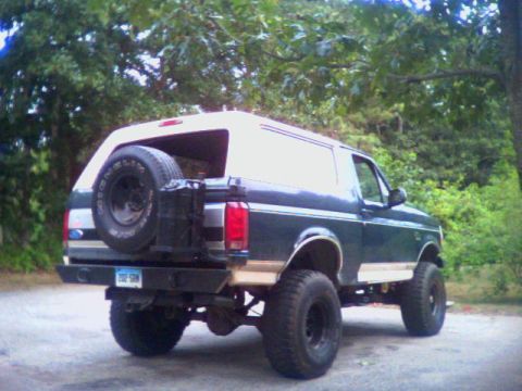 1993 Ford Bronco - Rear three-quarter view of a lifted two-tone dark green and tan Ford Bronco with a white removable hardtop, parked on gravel in front of he…