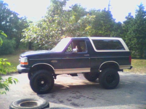 1993 Ford Bronco - Driver-side profile of a lifted dark green Ford Bronco with tan lower body cladding and a white removable hardtop, sitting on a paved drive…