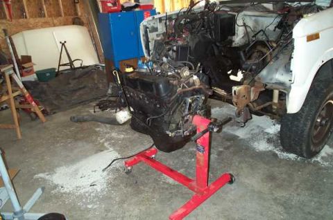 1985 Ford Bronco - An engine is mounted on a red stand in a garage, adjacent to a white vehicle undergoing engine work.