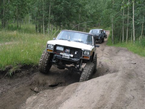 1981 Ford Bronco - Two Jeep Cherokee XJs navigate a challenging off-road trail through a wooded area.