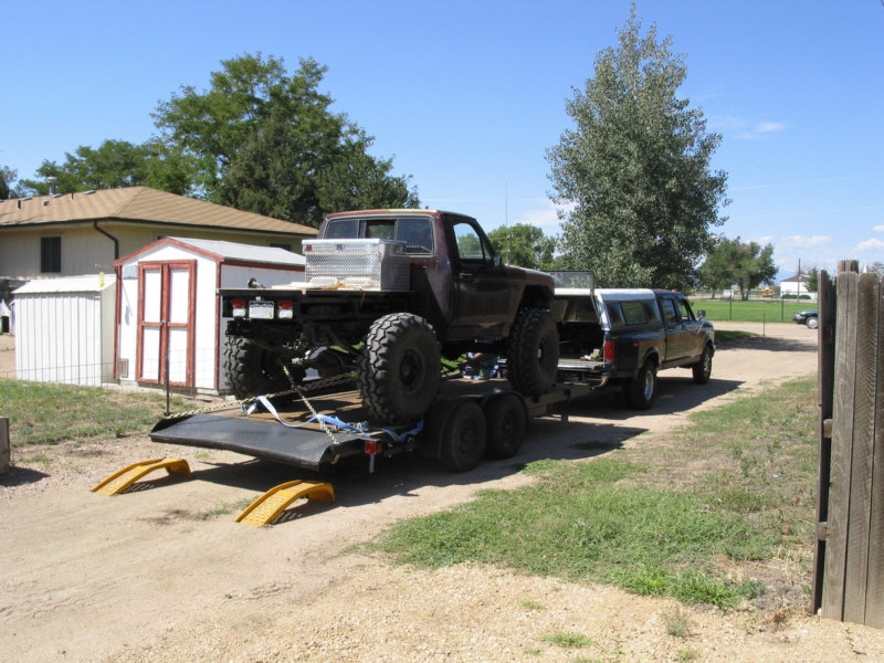 Maroon flatbed 4x4 on big tires strapped to a tandem trailer behind a crew cab pickup