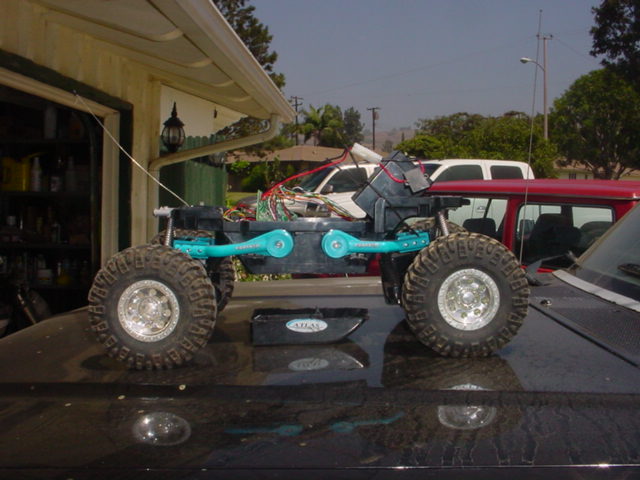 Scale RC rock crawler with teal chassis and chrome beadlock wheels posed on a black car hood