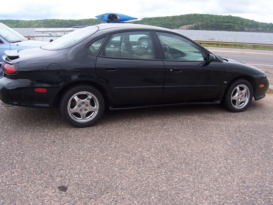 Black sedan parked by a lake, with a blue kayak on its roof. Another car is partially visible