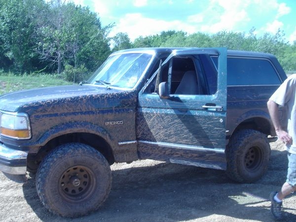 Mud-covered dark green Ford Bronco SUV with lift and large tires, side view on dirt with trees behind