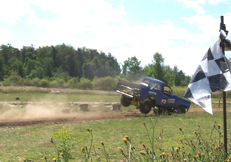 Blue lifted pickup airborne off a dirt jump near a black-and-white checkered flag