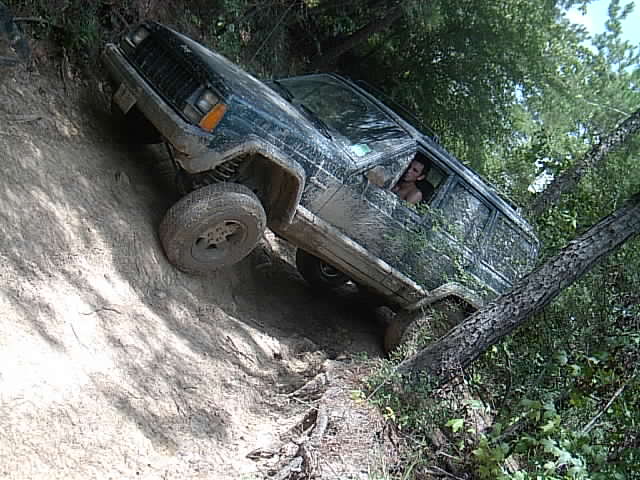 Mud-splattered Jeep Cherokee crawling up a steep dirt bank beside a tree, driver visible