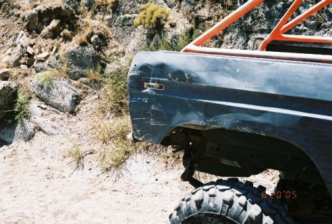 1979 Ford Bronco - Rear view of a damaged dark blue SUV with an orange roll cage, parked on a dirt and rock terrain.