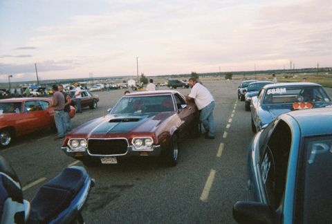 1972 Ford Ranchero - A man leans on a red 1972 Ford Ranchero at a drag strip with other vehicles in the background.