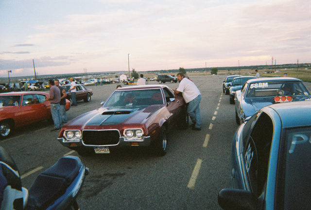 Maroon 1970s Ford with black hood stripes in a drag strip staging lane at dusk