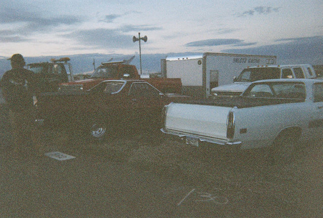 White car-based pickup and maroon sedan parked on a dirt lot at dusk