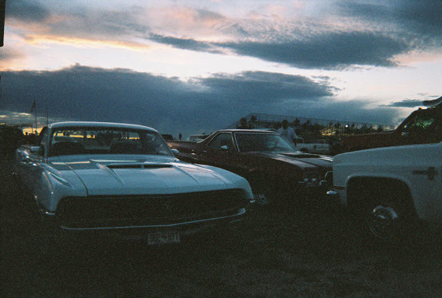 White Ford Ranchero with hood scoop parked in dirt lot at dusk beside other classics