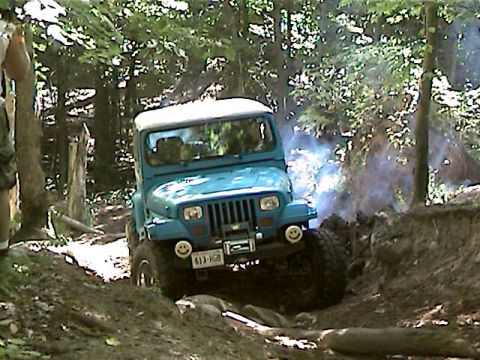 1977 Ford F-150 - A blue Jeep Wrangler navigates a challenging dirt path in a wooded area, with smoke billowing from its engine.