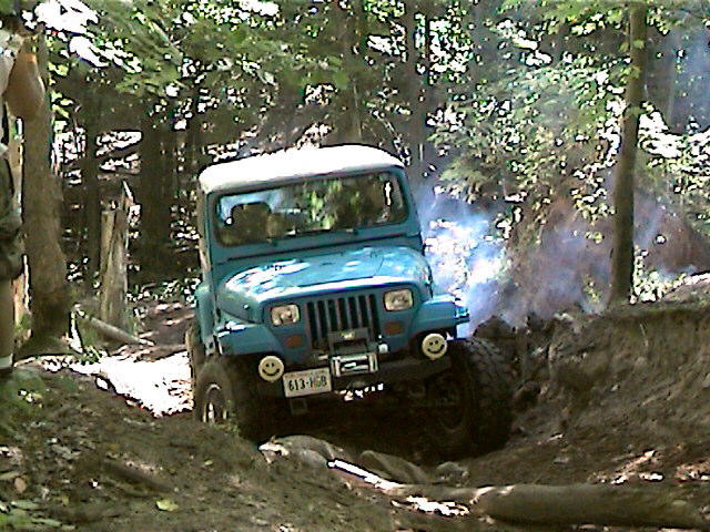 Blue Jeep Wrangler with a white top driving on a dirt trail, emitting smoke from the engine