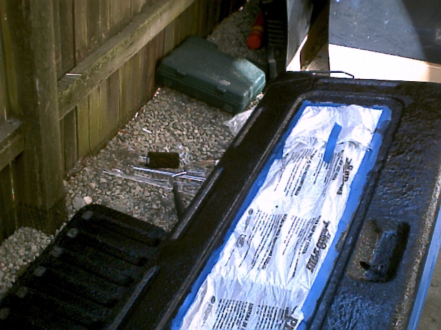 Black bedliner-coated tailgate face-up on gravel with new bedliner kit in plastic by a wood fence