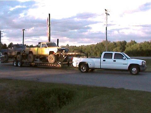 1977 Ford F-150 - A white pickup tows a trailer loaded with two other trucks under a cloudy sky.