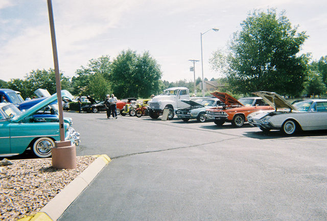 Several classic cars and a truck parked in a lot, some with hoods up. A turquoise car is in the foreground