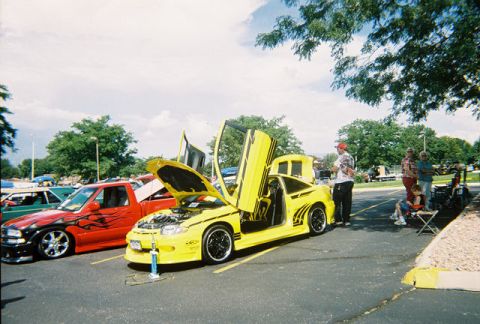1972 Ford Ranchero - A modified yellow car with butterfly doors open sits next to a red truck at an outdoor car show.