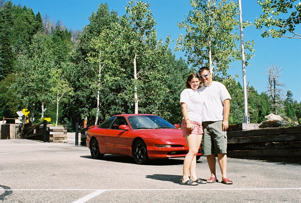 Couple standing in front of a red Ford Probe coupe in a tree-lined parking lot