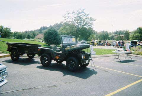 1972 Ford Ranchero - A military jeep and trailer are displayed at an outdoor car show with other vehicles in the background.