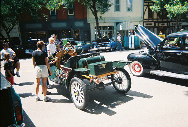 Green brass-era runabout with wooden spoke wheels at a downtown street car show