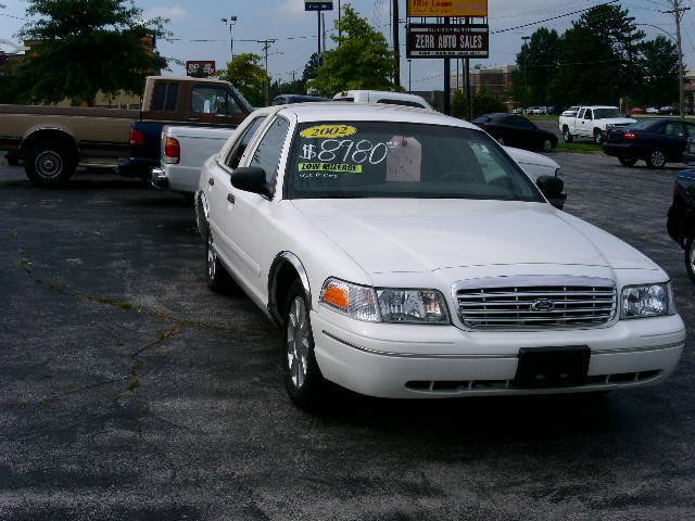 White Ford Crown Victoria with a "2002 $8780 LOW MILEAGE" sticker on the windshield, parked on asphalt