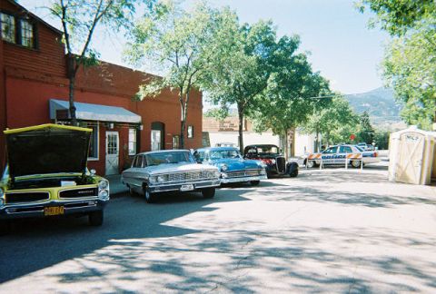 1972 Ford Ranchero - A collection of classic automobiles are parked along a street, with a police car and portable restrooms nearby under a clear sky.