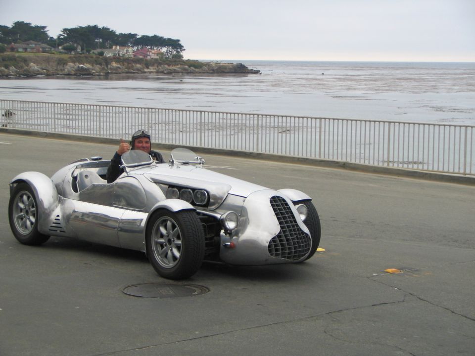 Polished bare-aluminum vintage-style open-wheel roadster on a coastal road, driver waving