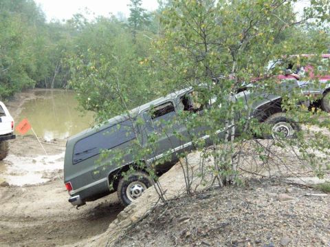 1995 Ford Bronco - A dark green square-body Chevrolet/GMC full-size SUV climbs a steep sandy embankment at a sharp nose-up angle beside a muddy water hole, se…