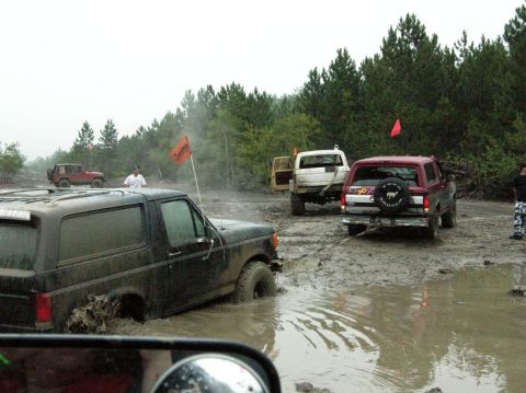 1995 Ford Bronco - Multiple off-road vehicles navigate a muddy terrain, with a black 1995 Ford Bronco prominently stuck in the mire.