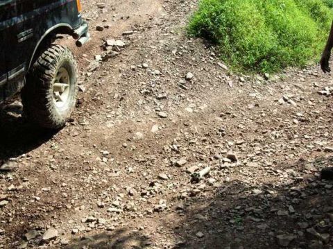 1995 Ford Bronco - Off-road vehicle driving on a rough, rocky trail with green vegetation to the right.