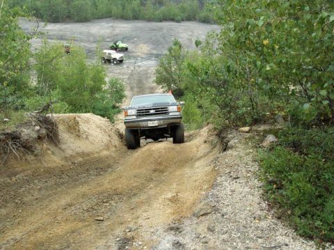 1995 Ford Bronco - Head-on view of a lifted 1980s-90s Ford truck/SUV on knobby mud tires climbing a rutted dirt hill on an off-road trail, front axle articula…