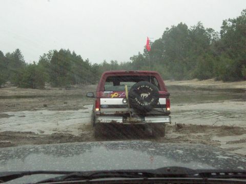 1995 Ford Bronco - A red Ford Bronco navigates a muddy trail, with a spare tire mounted on the back and a red flag visible.