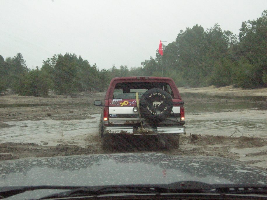 Red Ford Bronco driving through a muddy area, seen from another vehicle's perspective