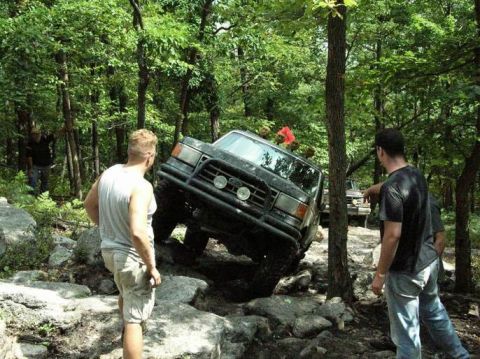 1995 Ford Bronco - A black 1995 Ford Bronco maneuvers over a challenging rocky terrain in a wooded area, with two men observing.