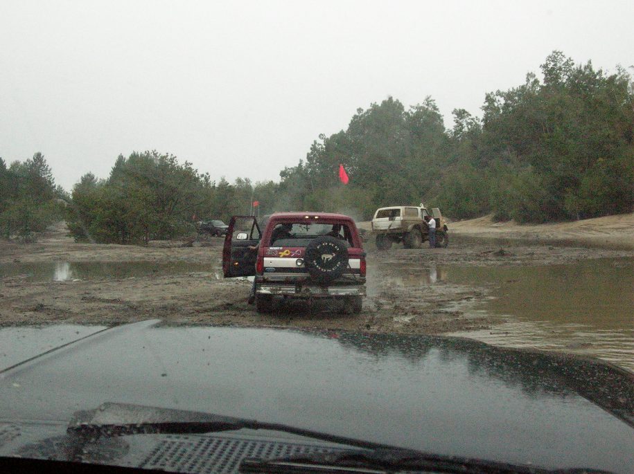 Red Ford Bronco with spare tire on rear, doors open, stuck in mud. Other vehicles visible in background