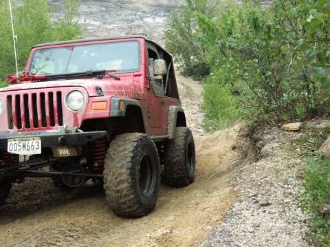 1995 Ford Bronco - A red Jeep Wrangler navigates a muddy, off-road trail surrounded by trees and rocky terrain.