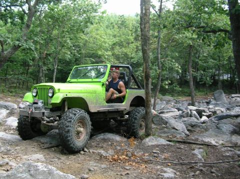 1995 Ford Bronco - A driver maneuvers a bright green Jeep through a challenging, rocky trail surrounded by trees.