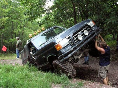1995 Ford Bronco - A dark blue lifted Ford truck tips hard onto its passenger side in a muddy wooded trail ditch, front wheel in the air, while a man in a bla…