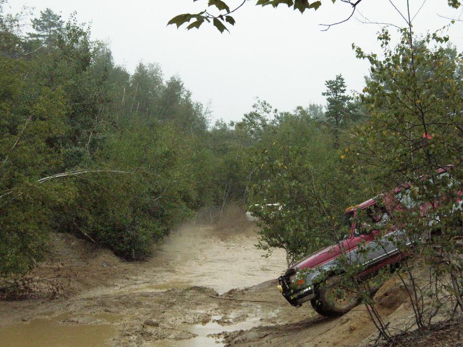 Red 1995 Ford Bronco driving through a muddy off-road trail with trees on either side