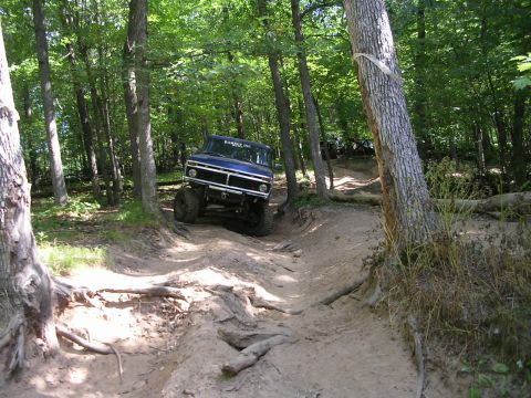 1977 Ford F-150 - A blue 1977 Ford F-150 navigates a challenging off-road trail through a wooded area, with other vehicles visible in the background.