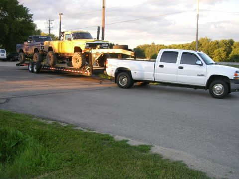 1977 Ford F-150 - A white dually pickup tows a trailer carrying two heavily modified trucks under a cloudy sky.