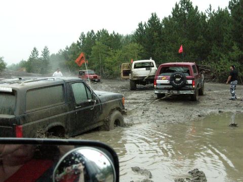 1995 Ford Bronco - Multiple off-road vehicles are stuck in a muddy terrain with trees in the background.