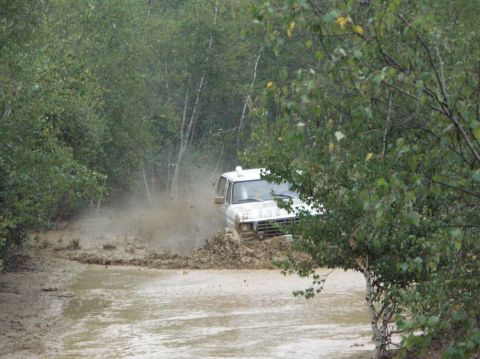 1995 Ford Bronco - A white 1980s Ford Bronco navigates a challenging muddy terrain, splashing water and dirt.