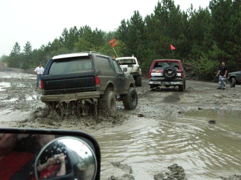 1995 Ford Bronco - Multiple off-road vehicles navigate a muddy terrain, kicking up water and mud.