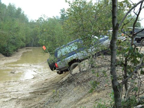 1995 Ford Bronco - A silver SUV navigates a challenging muddy trail with trees and water obstacles, while another vehicle is partially visible in the backgrou…