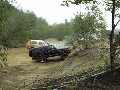 1995 Ford Bronco - Two SUVs tackle a challenging muddy off-road course with trees and foliage surrounding them.